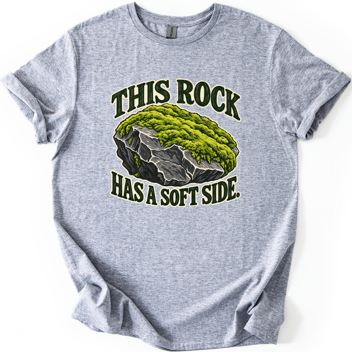 This Rock Has a Soft Side Comfort Colors T-shirt in Sport Grey, laid flat. A gray boulder topped with thick green moss sits between the words This Rock above and Has a Soft Side. below in dark green letters with a cream outline. The shirt lies on white with rolled sleeves.