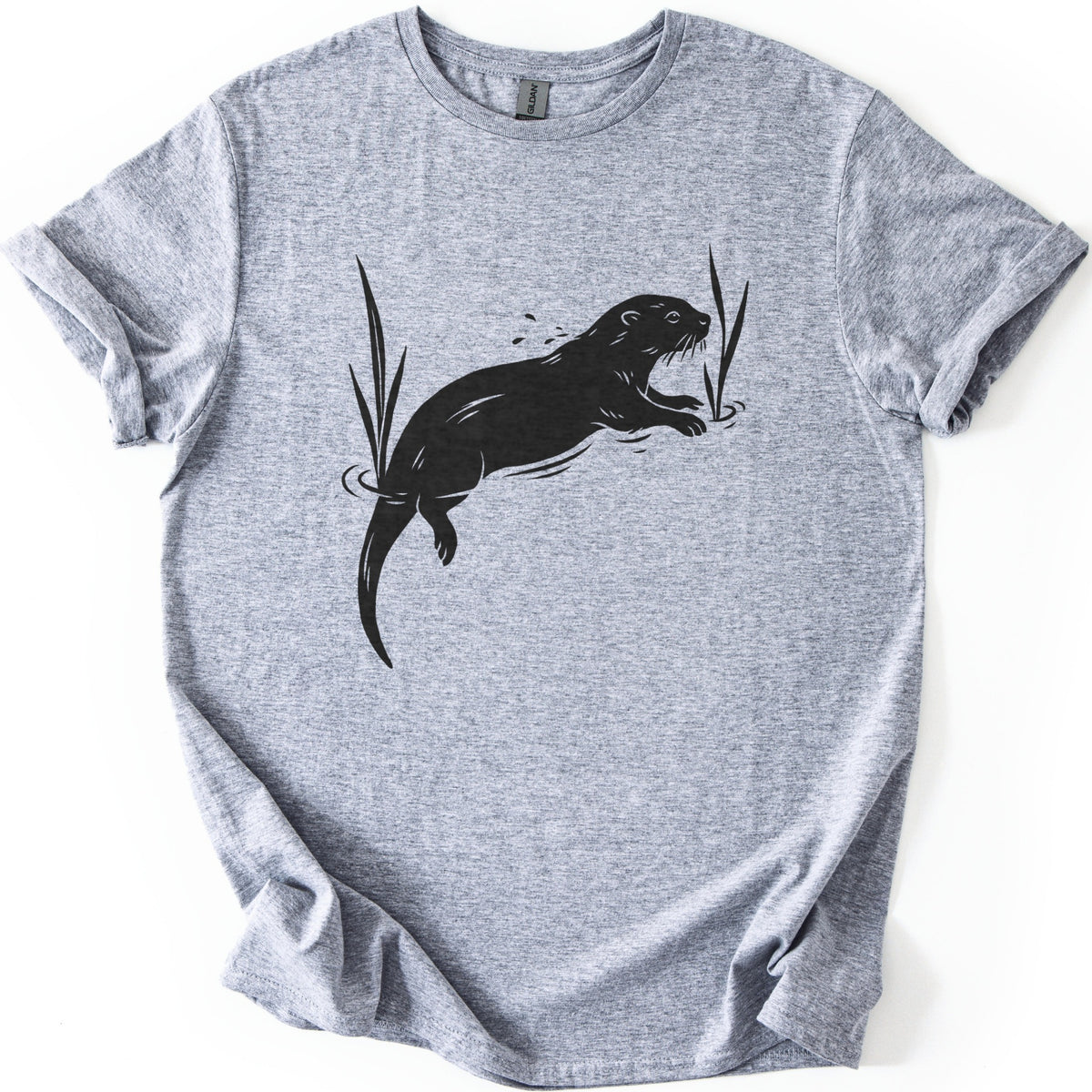 Swimming Otter Comfort Colors T-shirt in Sport Grey, laid flat. A black linocut-style otter swims through rippling water between tall reeds, with droplets above its head. No words are printed. The sleeves are cuffed and the shirt lies on a white background.