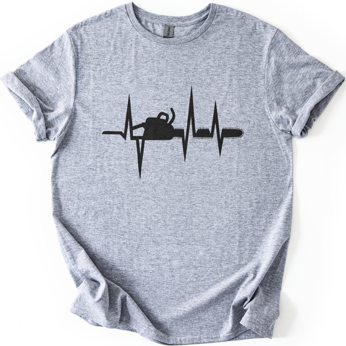 Chainsaw Heartbeat Comfort Colors T-shirt in Sport Grey, laid flat. A black heartbeat line with sharp peaks runs across the chest, with a chainsaw silhouette set into the line, its bar and chain extending to the right. The sleeves are rolled on a white background.