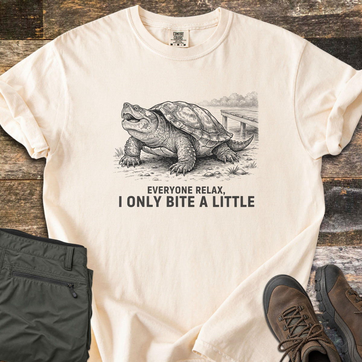 Everyone Relax I Only Bite A Little Comfort Colors T-shirt in Ivory, laid flat. A charcoal sketch of a snapping turtle with open mouth on a lakeshore by a wooden dock, above the words Everyone relax, I only bite a little. Olive hiking pants and brown hiking boots sit on a weathered wood floor.