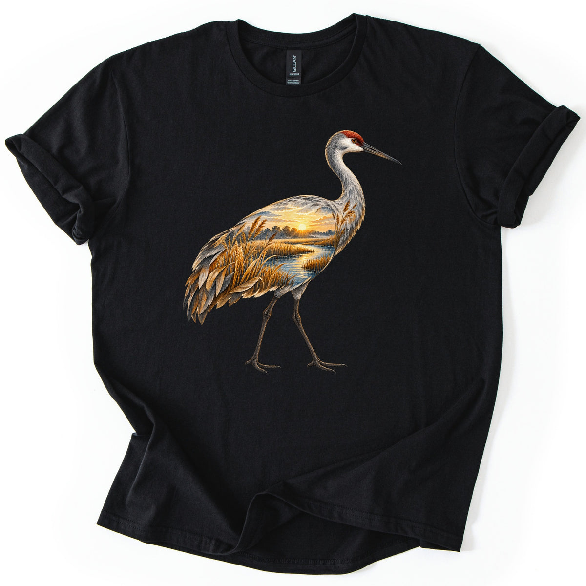 Crane Marsh Sunrise Comfort Colors T-shirt in Black, laid flat. A sandhill crane in mid-stride, red crown and gray neck, its body showing a sunrise marsh scene with golden reeds, a winding stream and treeline. No words. Cuffed sleeves on a white background.