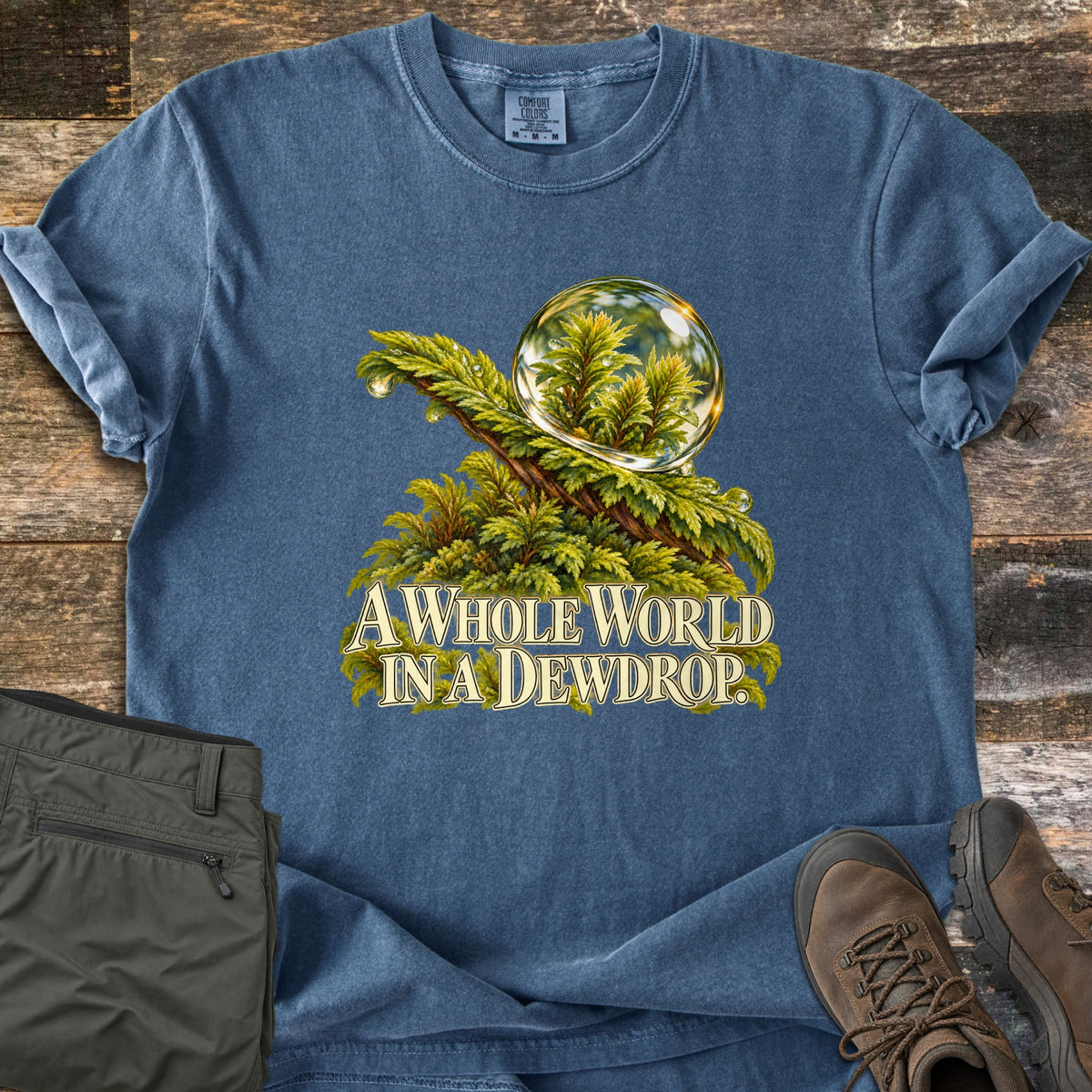A Whole World in a Dewdrop Comfort Colors T-shirt in Denim, laid flat. A clear dewdrop rests on a mossy fern branch, reflecting tiny green plants, above the words A Whole World in a Dewdrop. Olive hiking pants and brown hiking boots sit beside it on weathered wood planks.