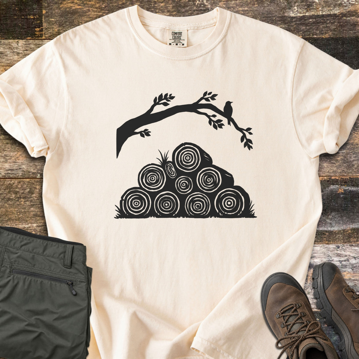 Stacked Firewood Comfort Colors T-shirt in Ivory, laid flat. A black print of a leafy tree branch with a small bird perched on it, arching over a pyramid of stacked firewood logs showing tree rings, with no words. Olive cargo pants and brown hiking boots sit on weathered wood planks.