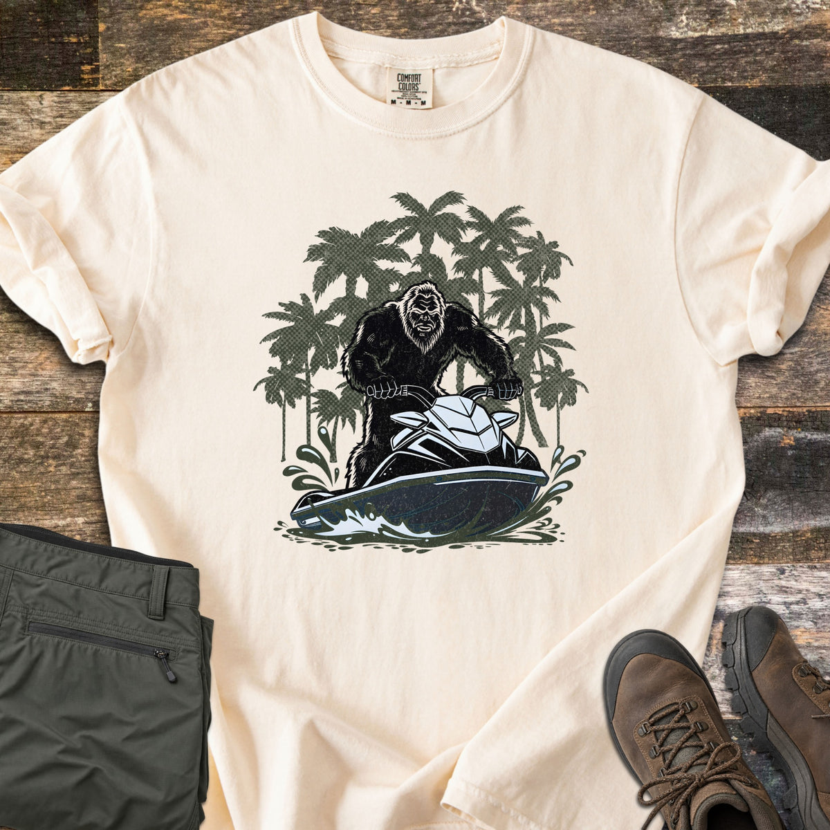 Jet Ski Squatch Comfort Colors T-shirt in Ivory, laid flat. Front print shows Bigfoot riding a black and white jet ski through splashing water, with dark green palm trees behind. Sleeves cuffed, on a wood surface with olive pants and brown hiking boots.