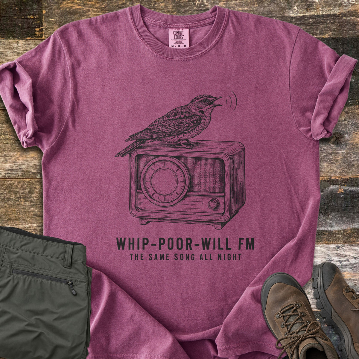 Whip-Poor-Will FM Comfort Colors T-shirt in Berry, laid flat. A sketched whip-poor-will sings atop a vintage radio above the words Whip-Poor-Will FM, The Same Song All Night. It lies with rolled sleeves on weathered wood beside folded olive pants and brown hiking boots.