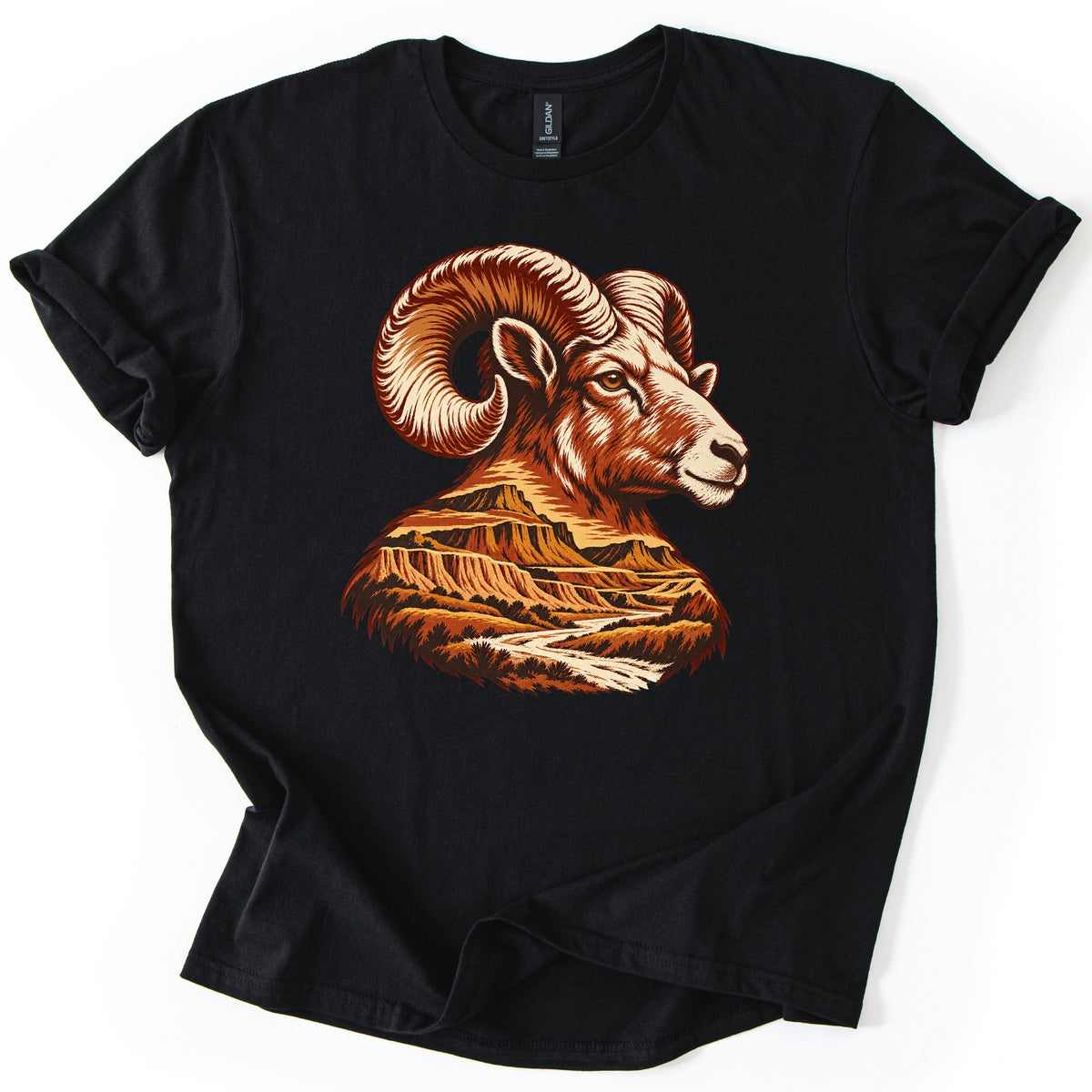 Bighorn Badlands Comfort Colors T-shirt in Black, laid flat. A rust and cream bighorn ram's head faces right, its curled horns above a neck that becomes a badlands scene of buttes and a winding river. No words are printed. The shirt lies on a white surface with rolled sleeves.