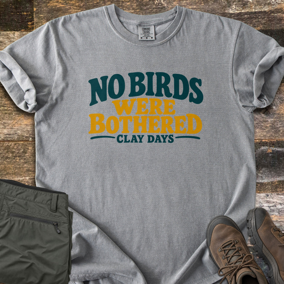 No Birds Were Bothered Comfort Colors T-shirt in Grey, laid flat. Printed on the chest in dark teal and mustard yellow lettering: No Birds Were Bothered, with Clay Days below. Sleeves rolled, on weathered wood with olive cargo pants and brown hiking boots.