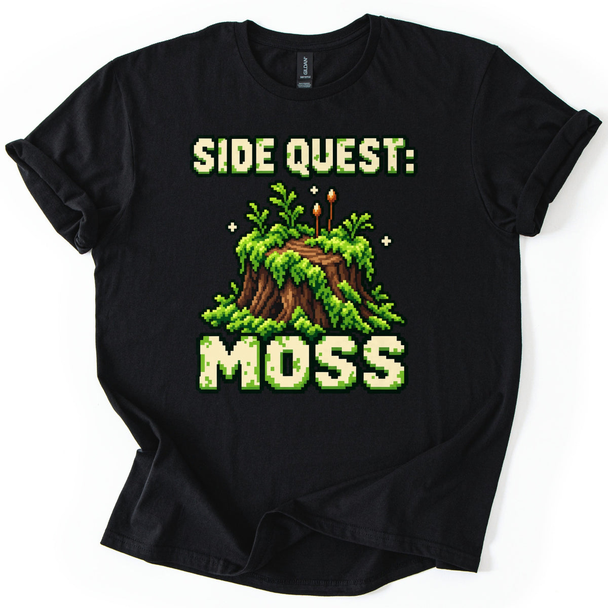 Side Quest Moss Comfort Colors T-shirt in Black, laid flat. Pixel-art tree stump draped in bright green moss and ferns with two small brown spore stalks, under the words Side Quest: and above Moss in mossy cream letters. Laid on a white surface with cuffed sleeves.