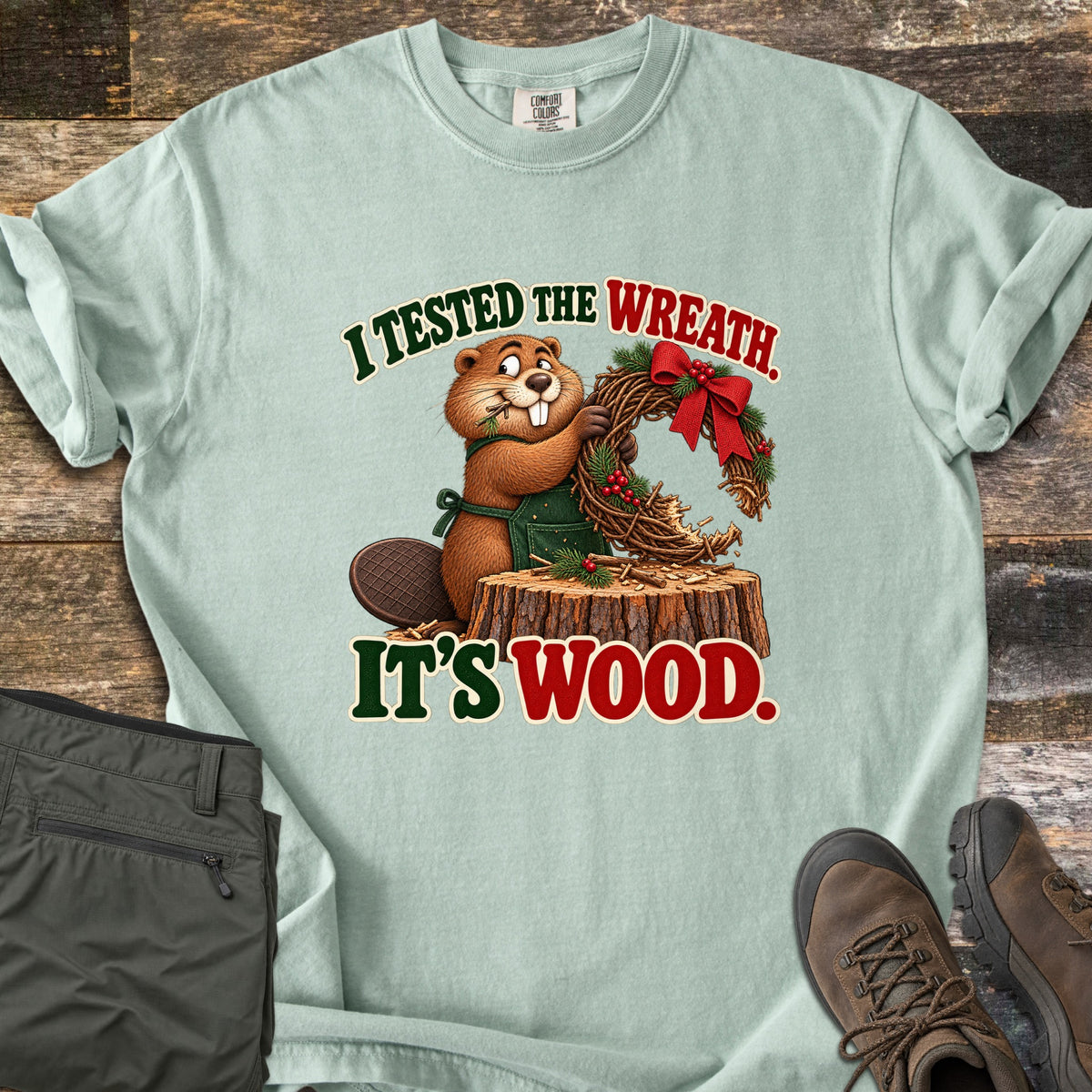 I Tested the Wreath. It's Wood. Comfort Colors T-shirt in Bay, laid flat. A cartoon beaver in a green apron chews a twig wreath with a red bow on a tree stump, under the words I Tested the Wreath. It's Wood. Olive pants and brown hiking boots lie beside it on weathered wood planks.