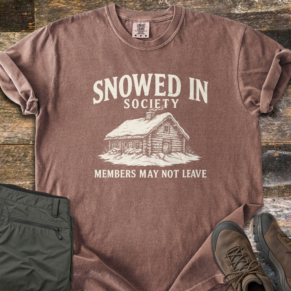 Snowed In Society Comfort Colors T-shirt in Espresso, laid flat. A cream print of a snow-covered log cabin with a smoking chimney sits under the words Snowed In Society and above Members May Not Leave. Folded olive pants and brown hiking boots lie beside it on weathered wood.