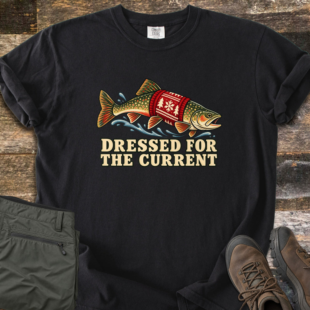 Dressed for the Current Comfort Colors T-shirt in Black, laid flat. A speckled trout wearing a red knit sweater with a white snowflake and tree pattern leaps through splashing water above the words Dressed for the Current. Olive pants and brown hiking boots lie on weathered wood.