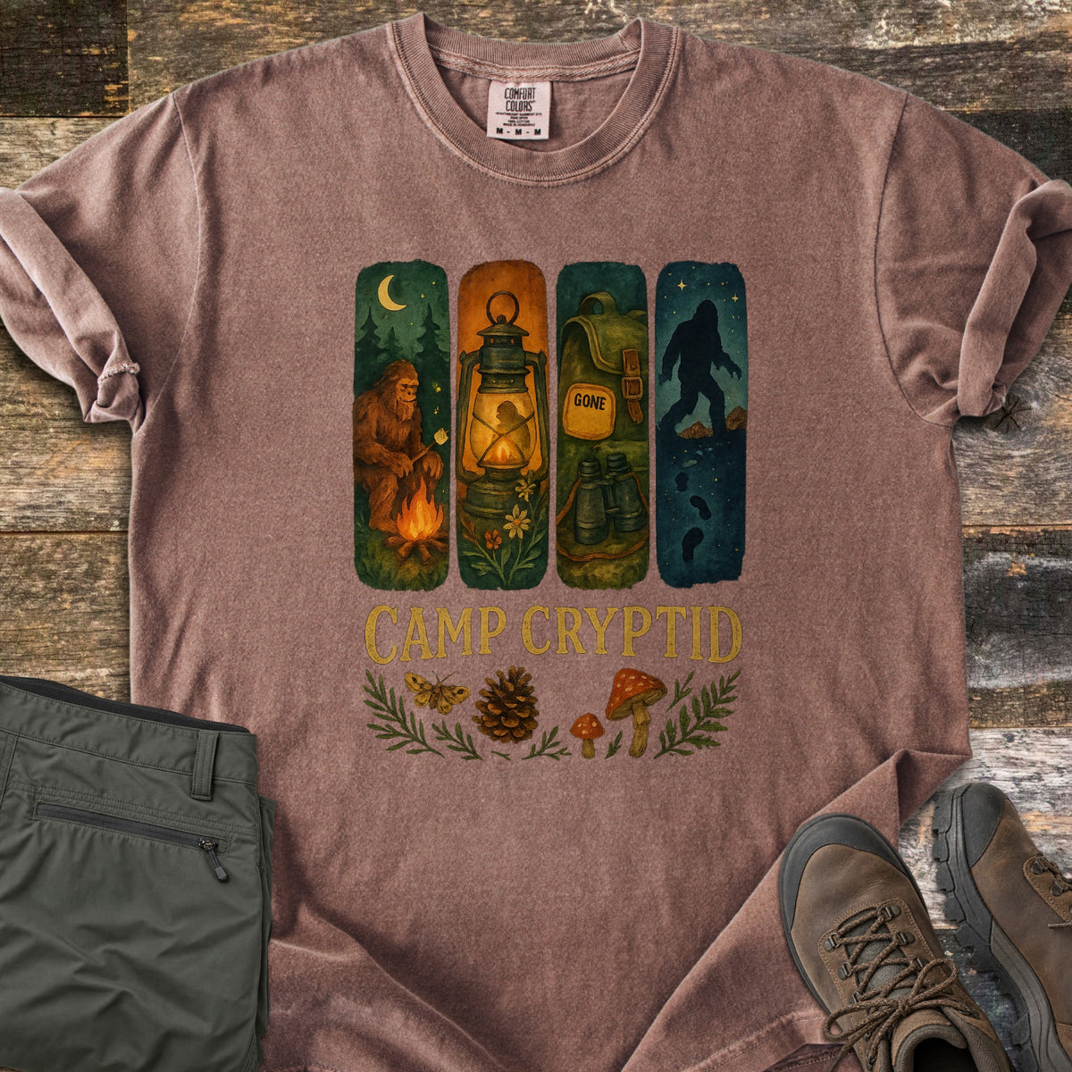 Camp Cryptid Big Foot Comfort Colors T-shirt in Espresso, laid flat. Four painted panels show Bigfoot at a campfire, a lantern, a backpack marked GONE with binoculars, and Bigfoot under stars, above CAMP CRYPTID, a moth, pinecone and mushrooms. Olive pants and hiking boots lie on wood.