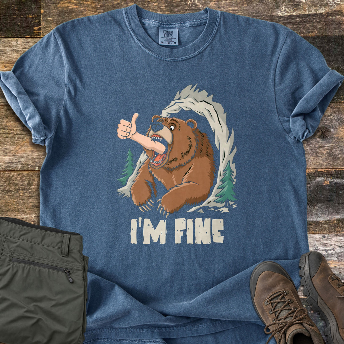 I'm Fine Comfort Colors T-shirt in Denim, laid flat. A cartoon brown bear bursts from a rocky cave framed by pine trees, a human arm in its mouth giving a thumbs-up, above the words I'M FINE. Olive pants and brown hiking boots sit on weathered wood.