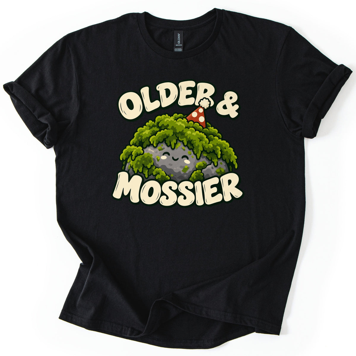 Older & Mossier Comfort Colors T-shirt in Black, laid flat. A smiling gray rock draped in green moss wears a red party hat with white dots, between the words Older & and Mossier in cream bubble letters. The shirt lies on a white background with its sleeves cuffed.
