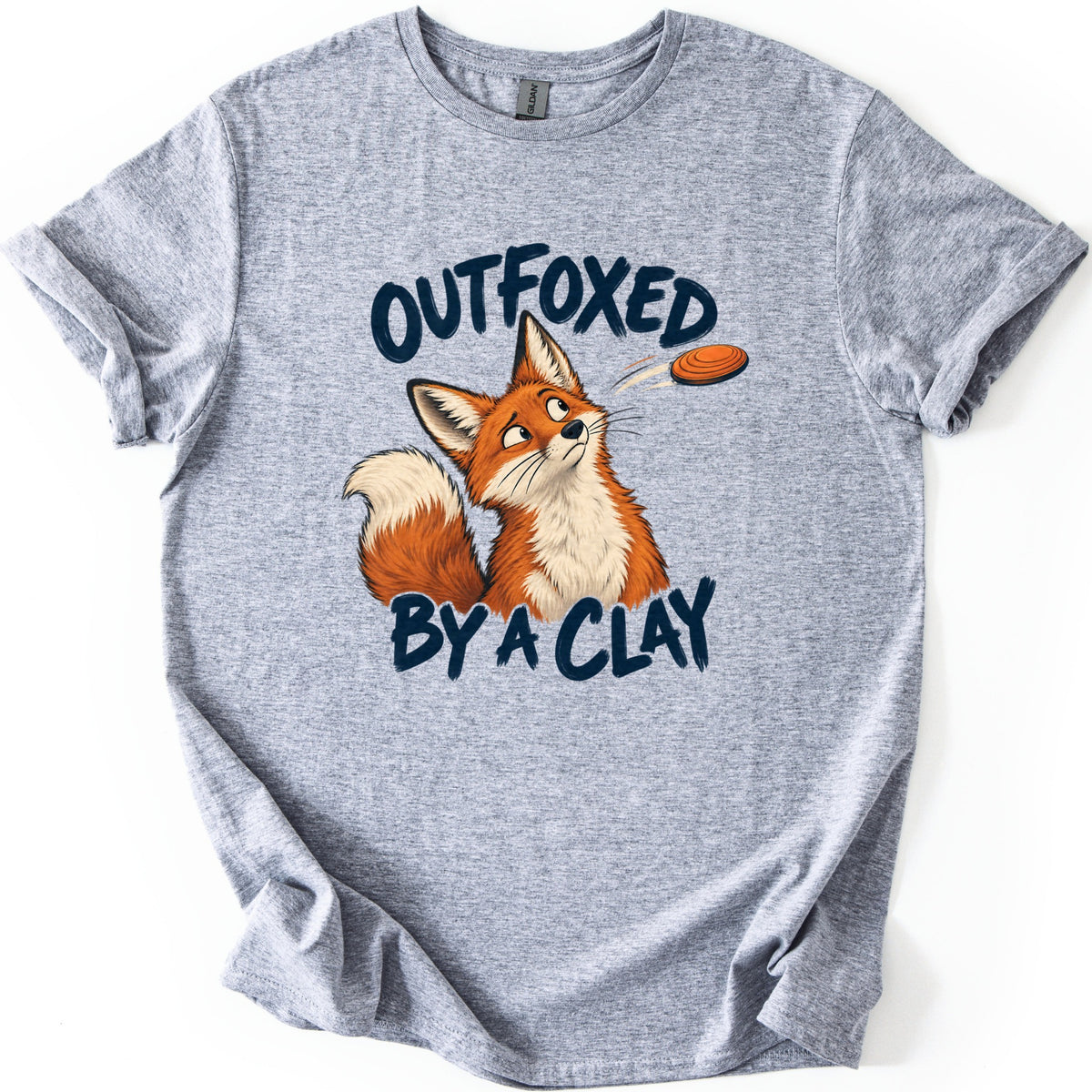 Outfoxed by a Clay Comfort Colors T-shirt in Sport Grey, laid flat. A cartoon red fox looks up warily at an orange clay target flying past, between the dark navy brush letters OUTFOXED and BY A CLAY. The shirt has rolled sleeves and lies on a white background.