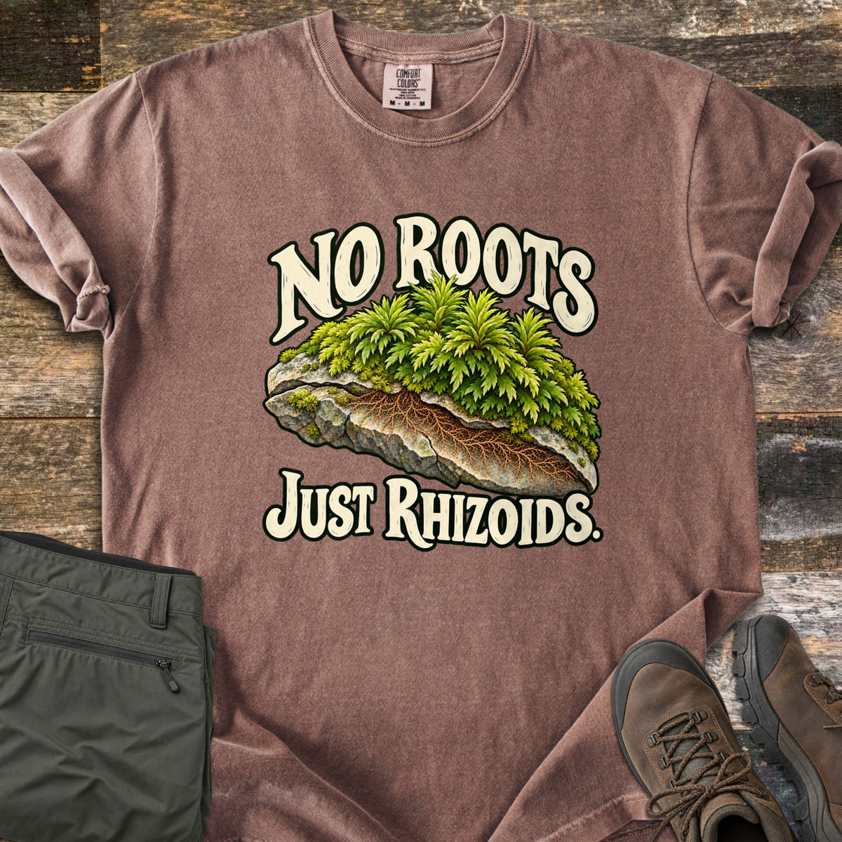 No Roots Just Rhizoids Comfort Colors T-shirt in Espresso, laid flat. Green moss sprouts from a cracked rock with brown rhizoids threading underneath, between the words No Roots Just Rhizoids. On rustic wood beside folded olive hiking pants and brown hiking boots.