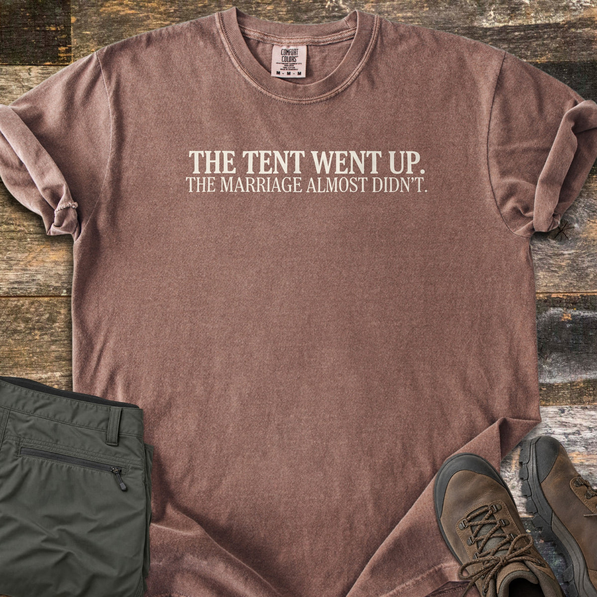 The Tent Went Up Comfort Colors T-shirt in Espresso, laid flat. Cream serif text across the chest reads The Tent Went Up. The Marriage Almost Didn't. Sleeves are rolled, with folded olive hiking pants and brown hiking boots beside it on weathered wood planks.