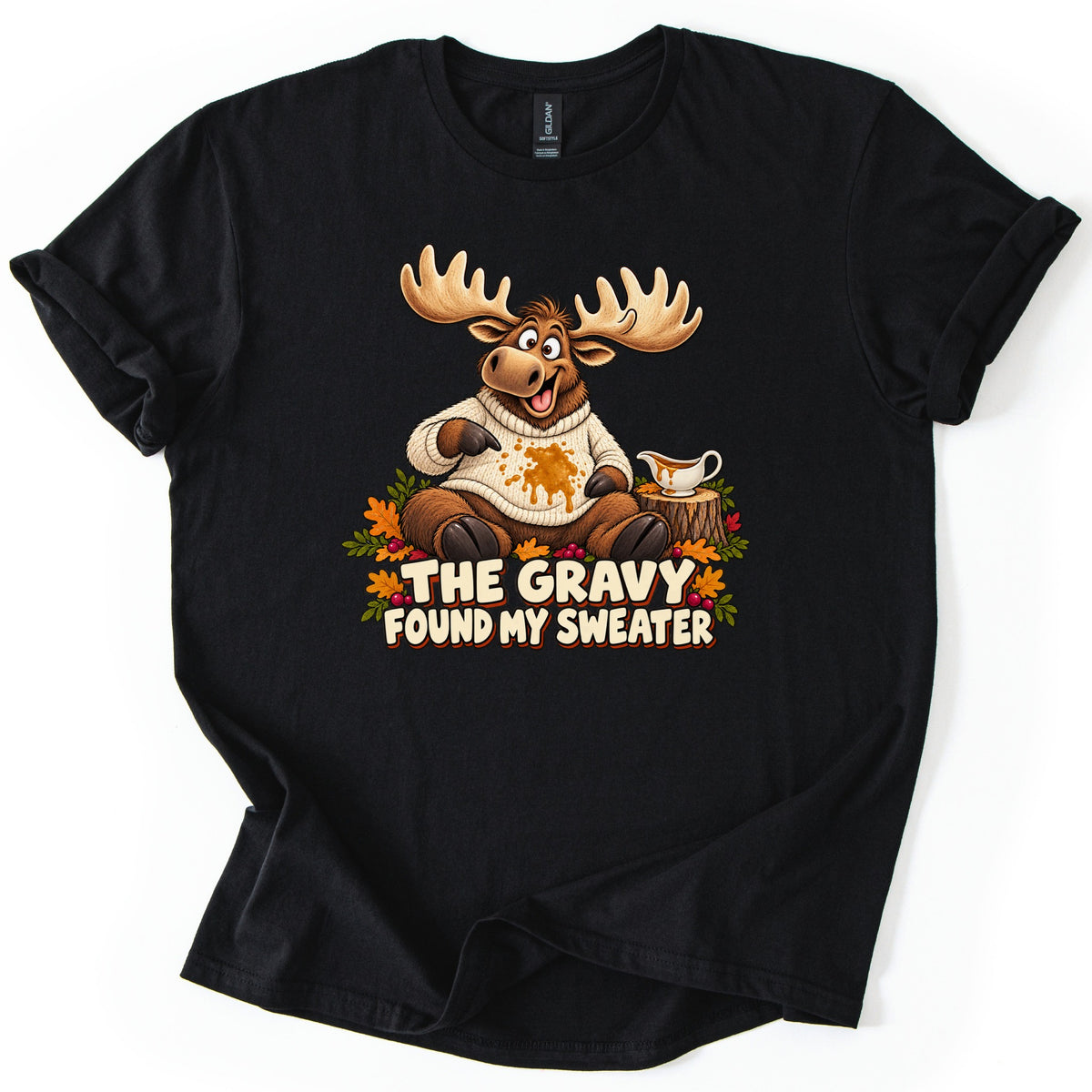 The Gravy Found My Sweater Comfort Colors T-shirt in Black, laid flat. A grinning cartoon moose in a cream knit sweater points at a gravy splash, next to a gravy boat on a stump with fall leaves and berries, above the words The Gravy Found My Sweater. Shown on a white background.
