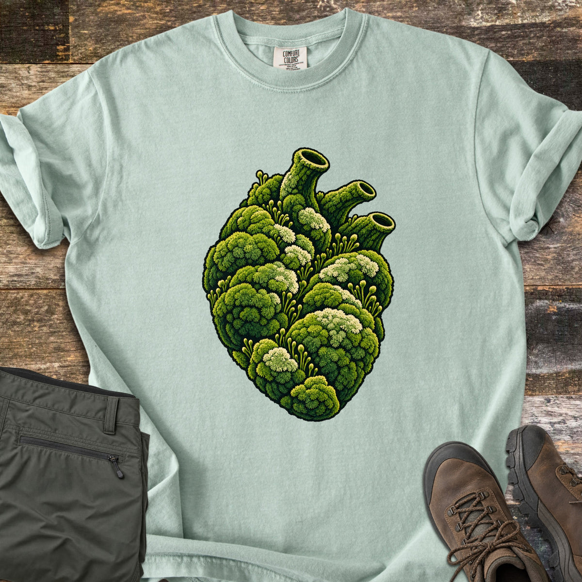 Moss Heart Comfort Colors T-shirt in Bay, laid flat. An anatomical heart covered in green moss with pale tufts, its vessels shaped like moss-covered tubes. The shirt has rolled sleeves and lies on weathered wood beside folded olive pants and brown hiking boots.