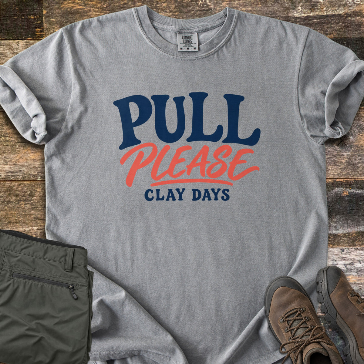 Pull Please Comfort Colors T-shirt in Grey, laid flat. The words Pull Please Clay Days are printed in navy and coral lettering, with Please in coral script. The shirt lies on weathered wood with rolled sleeves, beside folded olive pants and brown hiking boots.