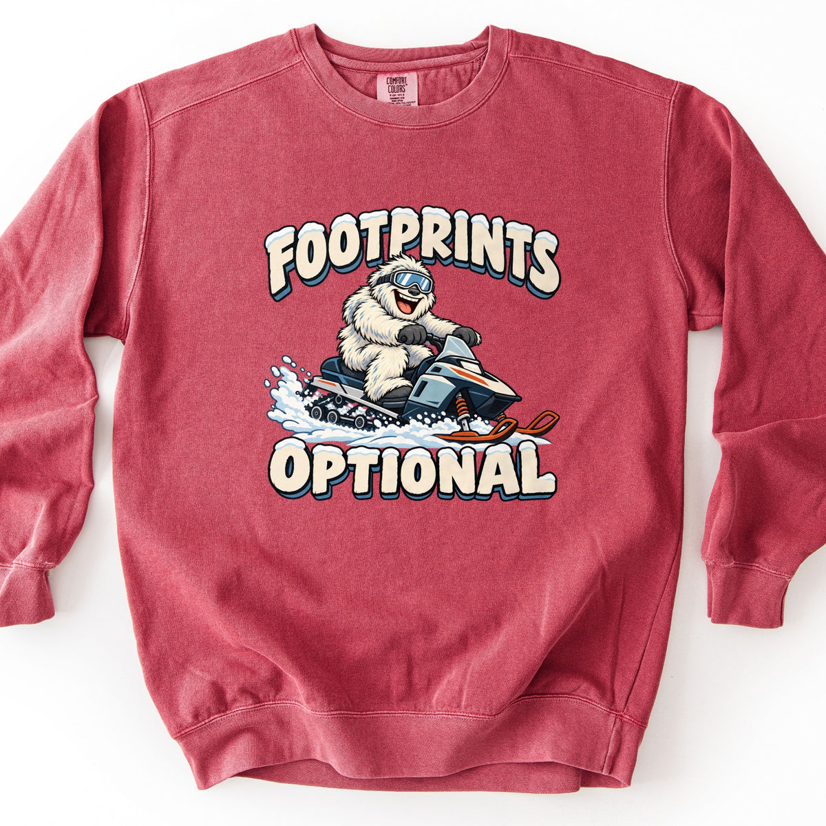 Footprints Optional Comfort Colors crewneck sweatshirt in Crimson, laid flat. A laughing furry white yeti wearing ski goggles drives a snowmobile kicking up snow, framed by snow-topped cream lettering that reads FOOTPRINTS above and OPTIONAL below. Shown on a white background.