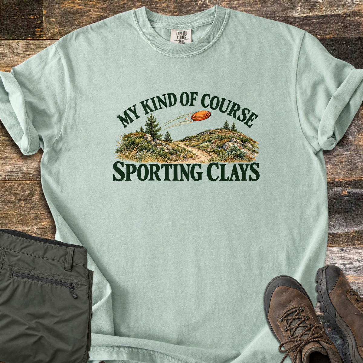 My Kind of Course Comfort Colors T-shirt in Bay, laid flat. The words My Kind of Course arch over a painted trail winding between grassy, rocky hills with pine trees as an orange clay target flies past, above Sporting Clays. Olive pants and brown hiking boots sit on weathered wood.
