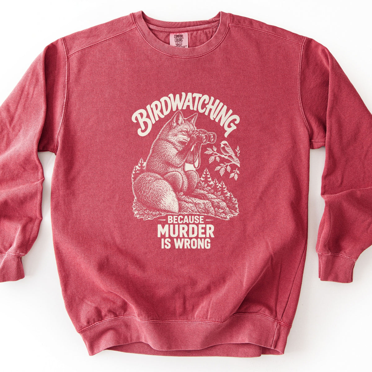 Birdwatching Because Murder Is Wrong Comfort Colors crewneck sweatshirt in Crimson, laid flat. A cream sketch of a fox on a rock using binoculars to watch a small bird on a leafy branch, with pine trees behind. Birdwatching arches above; Because Murder Is Wrong is below. White background.