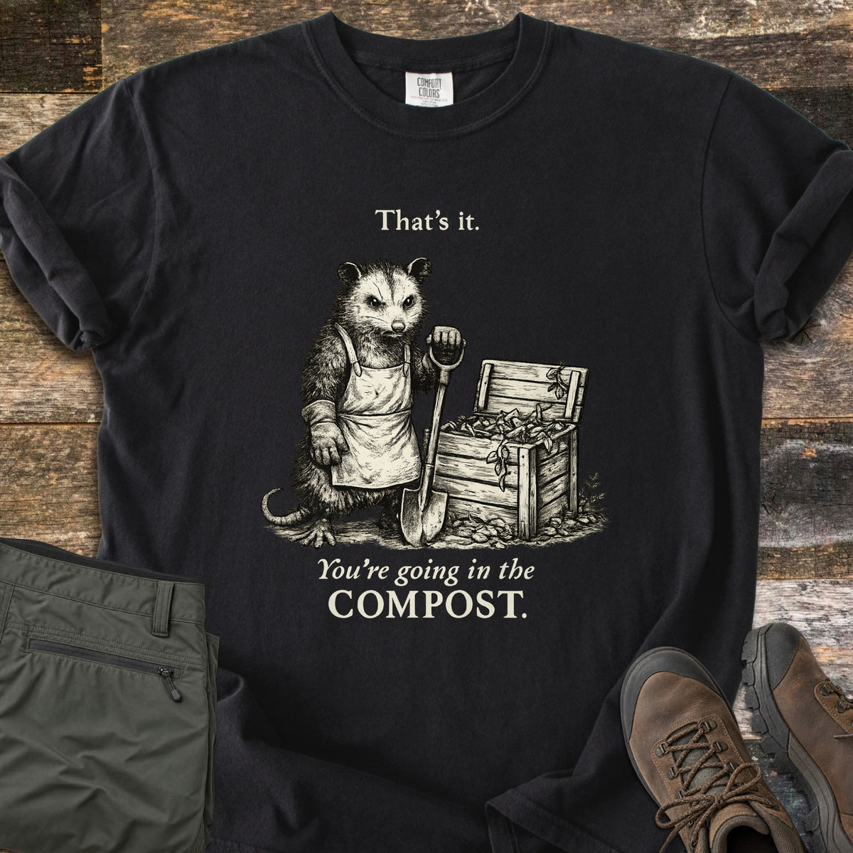 Going In The Compost Possum Comfort Colors T-shirt in Black, laid flat. Cream etching of a scowling possum in an apron, leaning on a shovel by a wooden compost bin, between the words That's it. and You're going in the COMPOST. Olive pants and brown boots lie on wood planks.