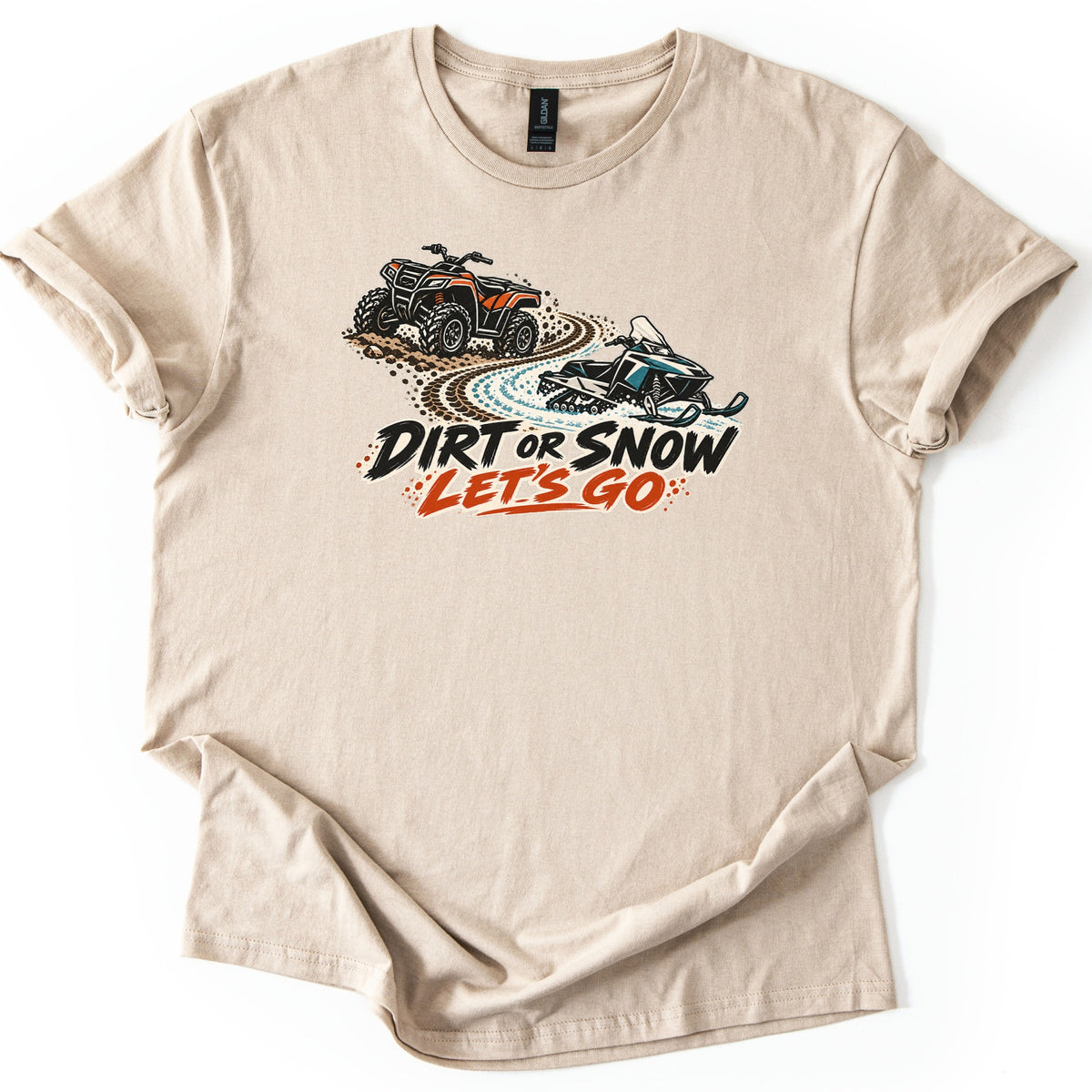 Dirt or Snow Let's Go Comfort Colors T-shirt in Sand, laid flat. An orange ATV kicks up dirt on a trail that turns into snow beneath a teal snowmobile, above the words DIRT OR SNOW LET'S GO. The shirt lies on a white background with cuffed sleeves.