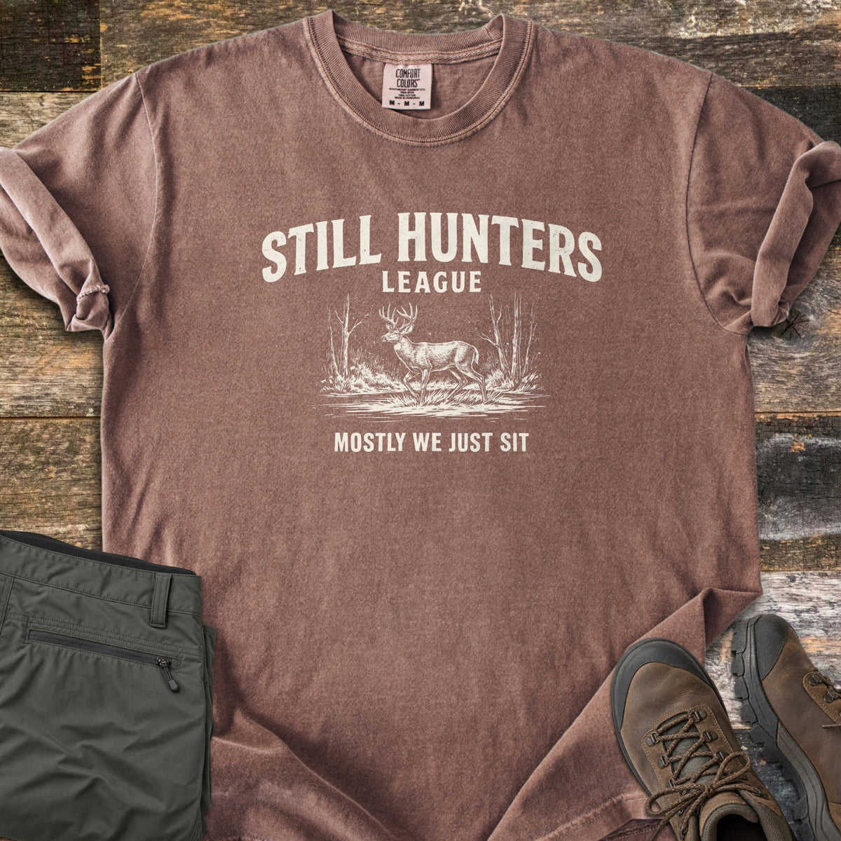 Still Hunters League Comfort Colors T-shirt in Espresso, laid flat. Cream print of the words Still Hunters League above a sketched buck deer standing in grass between bare trees, with Mostly We Just Sit below. Sleeves rolled, on weathered wood with olive pants and brown hiking boots.
