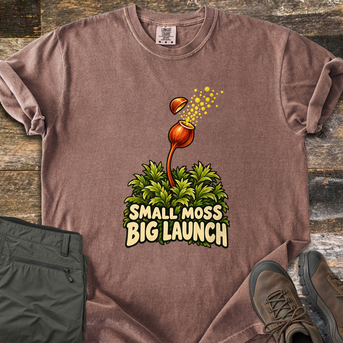 Small Moss Big Launch Comfort Colors T-shirt in Espresso, laid flat. A moss capsule on a thin orange stalk flips its lid off as yellow spores spray out above leafy green moss and the words Small Moss Big Launch. Folded olive pants and brown hiking boots rest on rustic wood.