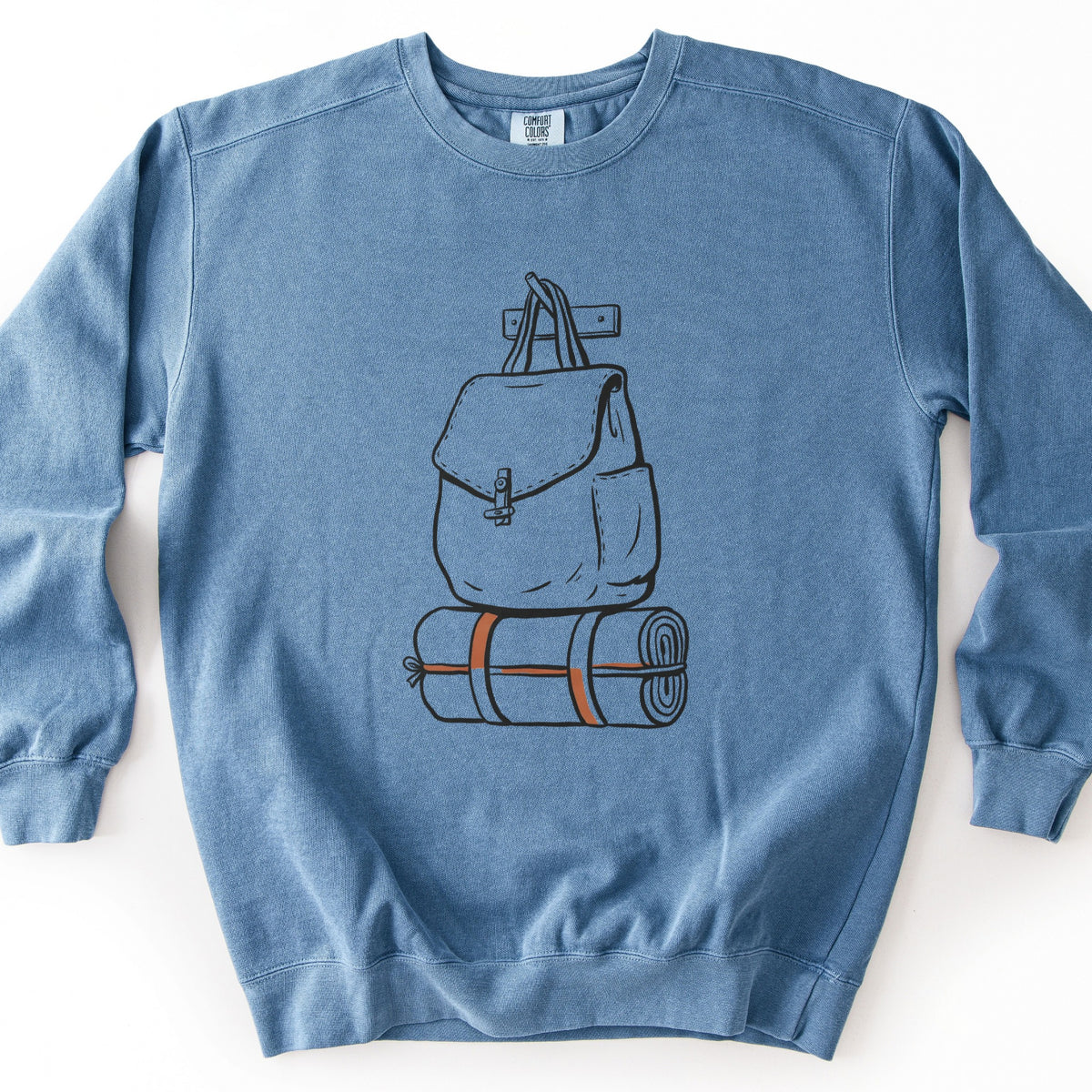 Backpack And Bedroll Comfort Colors crewneck sweatshirt in Blue Jean, laid flat. Black line art of a canvas backpack with a toggle clasp hanging by its straps from a wall peg above a rolled bedroll tied with orange straps. No words are printed. Shown on a white background.