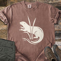 Otter In The Reeds Comfort Colors T-shirt in Espresso, laid flat. A cream silhouette of an otter with open mouth and raised paws curls behind two tall reeds above wavy water lines, no words. Folded olive hiking pants and brown hiking boots lie on weathered wood.