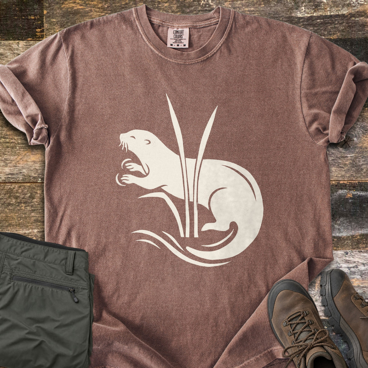 Otter In The Reeds Comfort Colors T-shirt in Espresso, laid flat. A cream silhouette of an otter with open mouth and raised paws curls behind two tall reeds above wavy water lines, no words. Folded olive hiking pants and brown hiking boots lie on weathered wood.