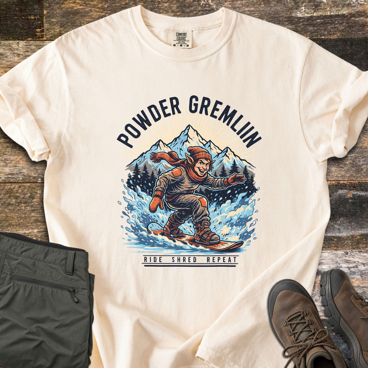 Powder Gremlin Comfort Colors T-shirt in Ivory, laid flat. Under the words Powder Gremlin, a grinning gremlin in a knit hat and orange scarf snowboards through blue powder below snowy peaks, above Ride Shred Repeat. Olive pants and brown hiking boots lie on weathered wood.