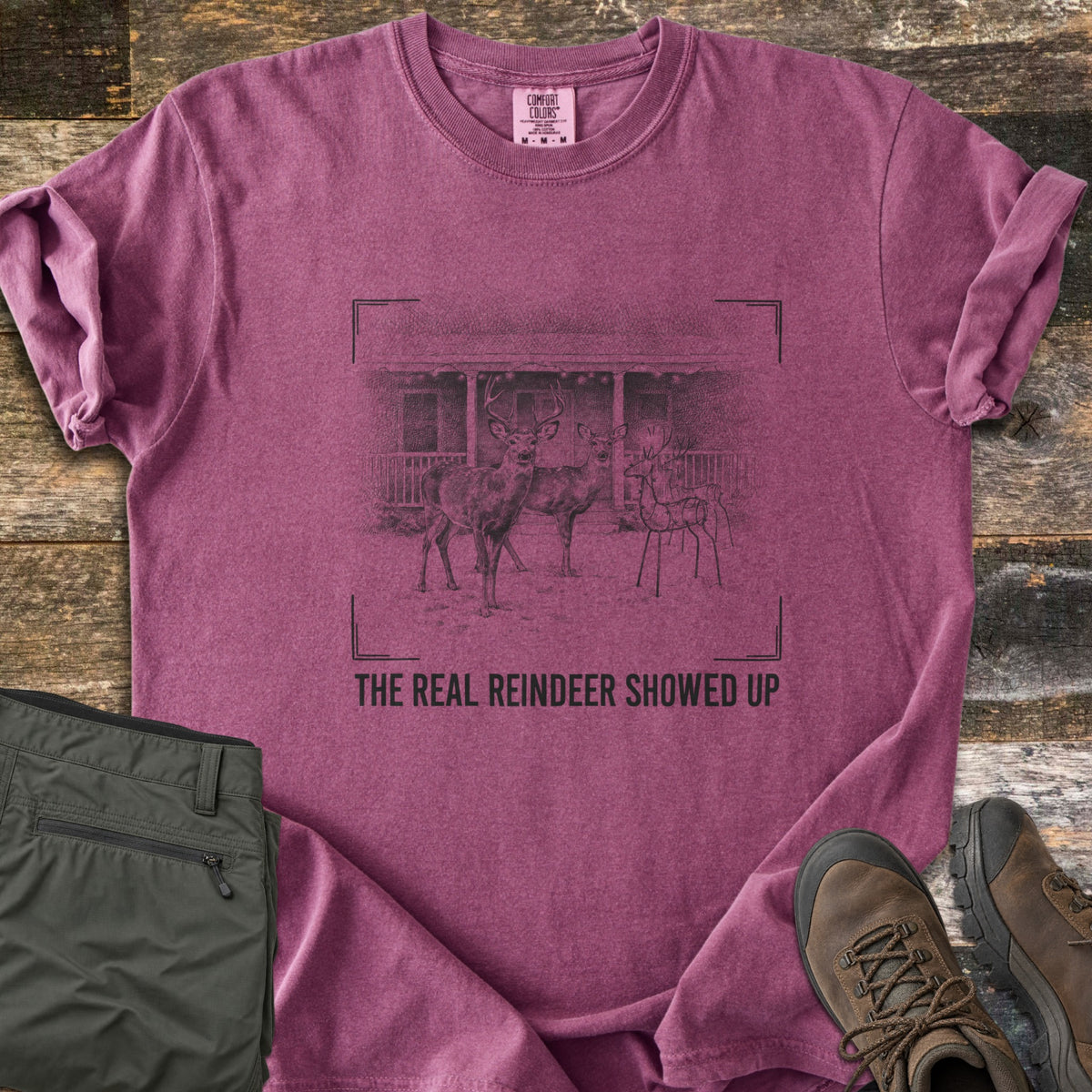 The Real Reindeer Showed Up Comfort Colors T-shirt in Berry, laid flat. A pencil-style sketch framed by viewfinder corners shows two real deer beside a wire-frame reindeer decoration at a lit porch, above the words The Real Reindeer Showed Up. Olive pants and hiking boots sit nearby.