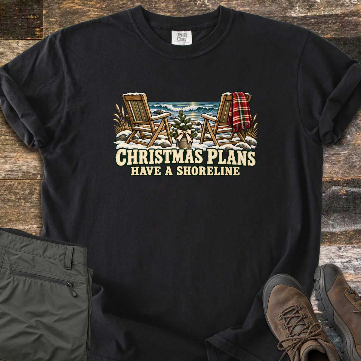 Christmas Plans Have a Shoreline Comfort Colors T-shirt in Black, laid flat. Two snow-dusted wooden Adirondack chairs, one with a red plaid blanket, flank a small potted tree with a bow before ocean waves, above the words Christmas Plans Have a Shoreline. Olive pants and brown boots lie on weathered wood.