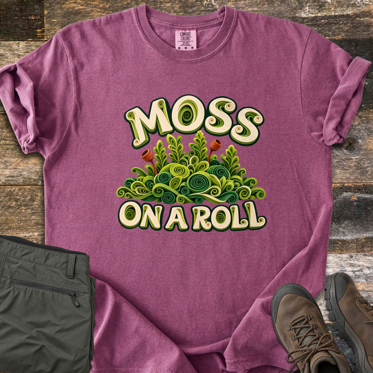 Moss on a Roll Comfort Colors T-shirt in Berry, laid flat. Swirled paper-quill style green moss with two rust-colored sprouts sits between the words Moss On A Roll in cream lettering. Sleeves are cuffed on a wood plank floor beside olive hiking pants and brown hiking boots.