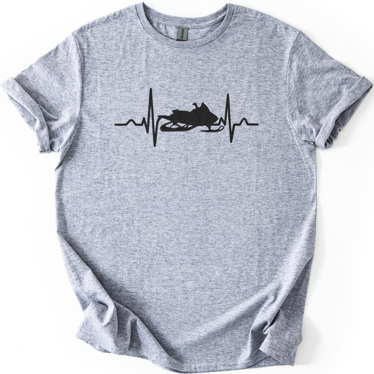 Heartbeat Snowmobile Comfort Colors T-shirt in Sport Grey, laid flat. A black heartbeat line crosses the chest, with a snowmobile silhouette in the center of the pulse and no words. The shirt has cuffed sleeves and lies on a white background.