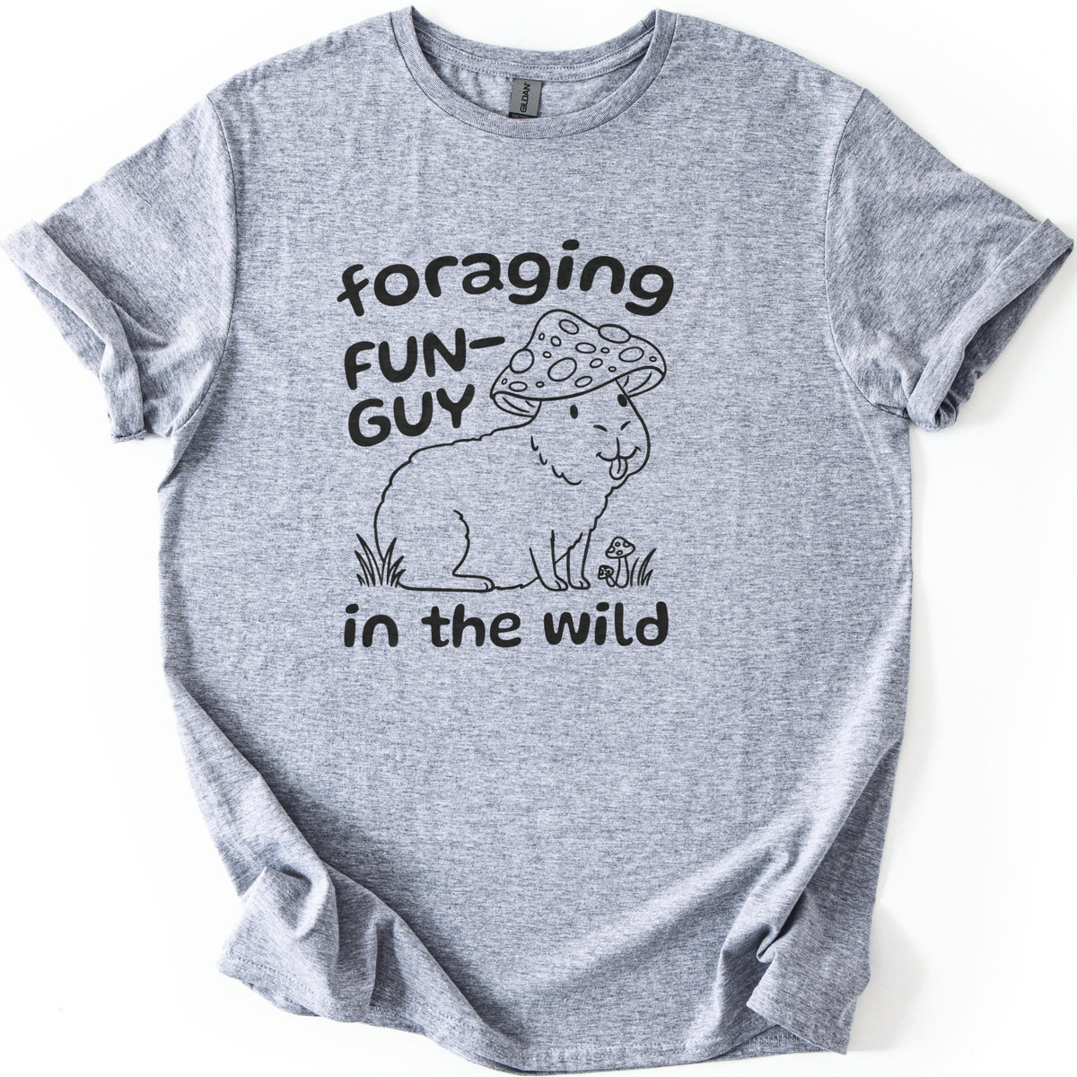 Foraging Fun-Guy Capybara Comfort Colors T-shirt in Sport Grey, laid flat. A black line drawing of a winking capybara with its tongue out, wearing a spotted mushroom cap and sitting in grass by tiny mushrooms, with the words foraging FUN-GUY in the wild. Sleeves rolled, on white.