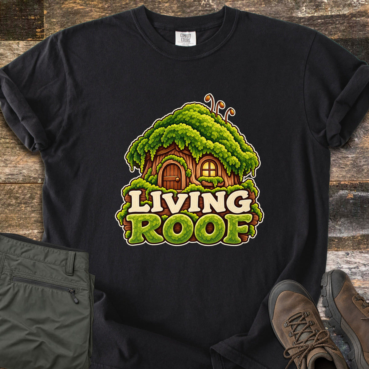 Living Roof Comfort Colors T-shirt in Black, laid flat. A cartoon tree-stump cottage with a wooden door, glowing window, and thick green moss roof sits above the words Living Roof in cream and mossy green letters. Olive hiking pants and brown hiking boots lie on weathered wood.