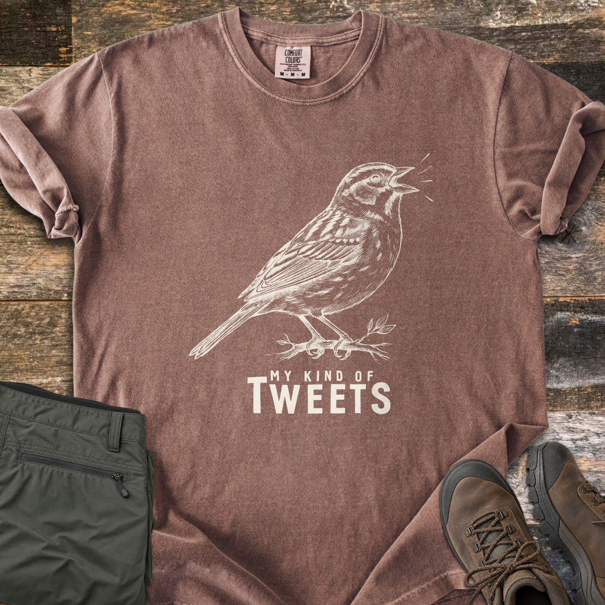 My Kind Of Tweets Comfort Colors T-shirt in Espresso, laid flat. A cream sketch of a singing sparrow perched on a leafy twig, above the words My Kind Of Tweets. Shown on rustic wood with folded olive hiking pants and brown hiking boots.