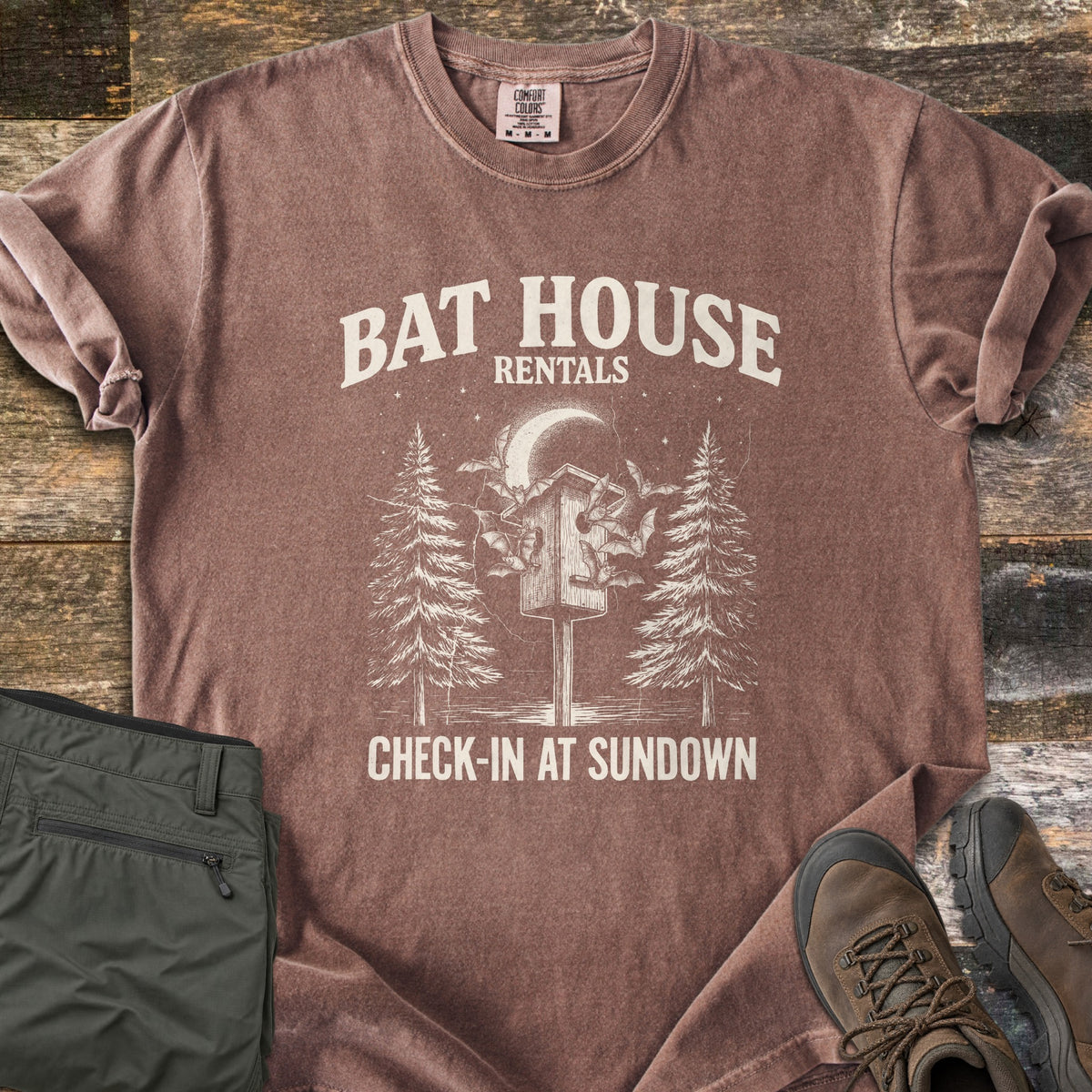 Bat House Rentals Comfort Colors T-shirt in Espresso, laid flat. Off-white print of a bat house on a post with bats flying around it under a crescent moon, between two pine trees, with the words Bat House Rentals and Check-In at Sundown. Olive pants and brown hiking boots lie on wood planks.