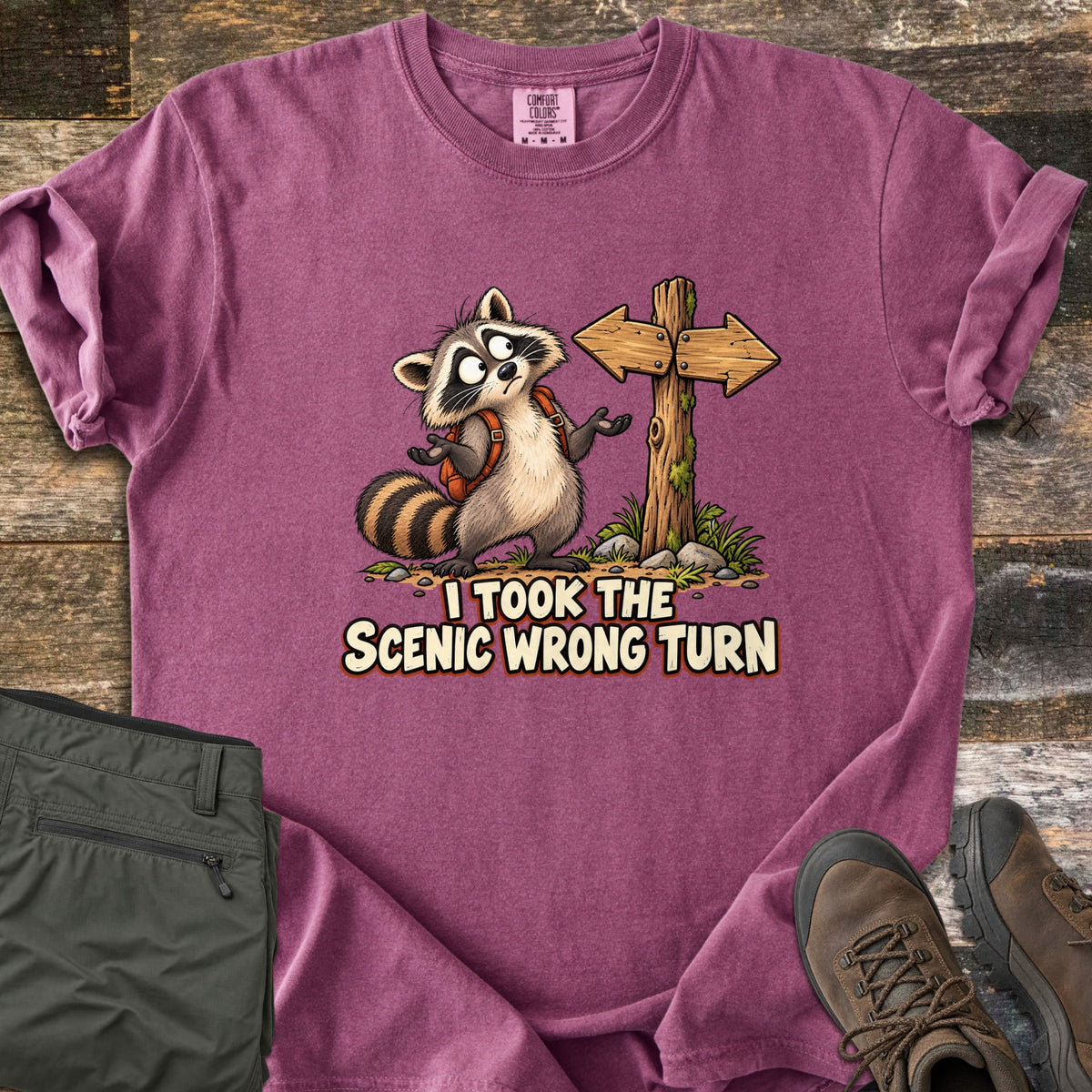 I Took the Scenic Wrong Turn Comfort Colors T-shirt in Berry, laid flat. A cartoon raccoon with a backpack shrugs, looking confused beside a wooden signpost with arrows pointing both ways, above the words I Took the Scenic Wrong Turn. Olive hiking pants and brown boots lie on weathered wood.