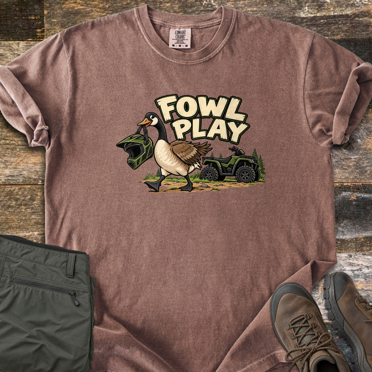 Fowl Play Comfort Colors T-shirt in Espresso, laid flat. A cartoon Canada goose carries a green off-road helmet in its beak past a green ATV and pine trees, under the words Fowl Play. Sleeves rolled, on weathered wood with olive pants and brown hiking boots.