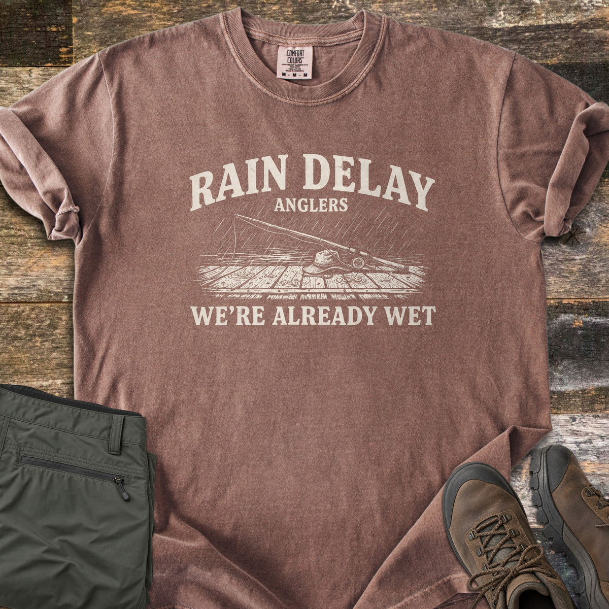 Rain Delay Anglers Comfort Colors T-shirt in Espresso, laid flat. Cream print of a fishing rod and bucket hat on a rain-soaked wooden dock, under the words Rain Delay Anglers and above We're Already Wet. Sleeves rolled, on rustic wood beside folded olive pants and brown boots.