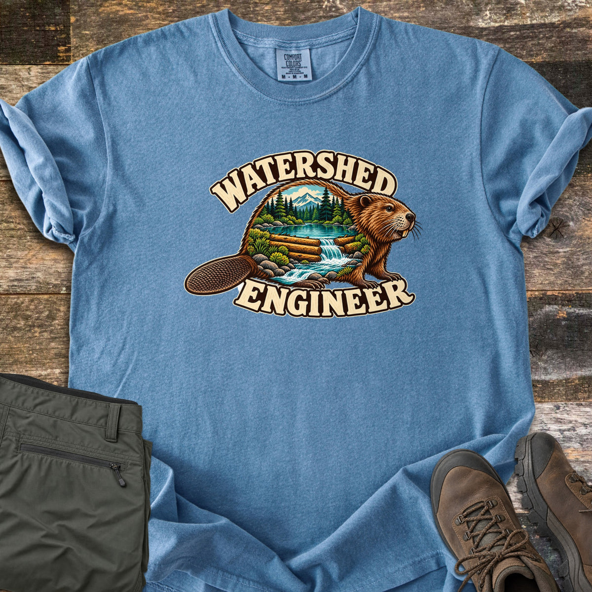 Watershed Engineer Comfort Colors T-shirt in Blue Jean, laid flat. A beaver whose body frames a forest scene with mountains, a lake, and a waterfall flowing over a log dam, between the words Watershed Engineer. Olive pants and brown hiking boots lie on a wood surface.