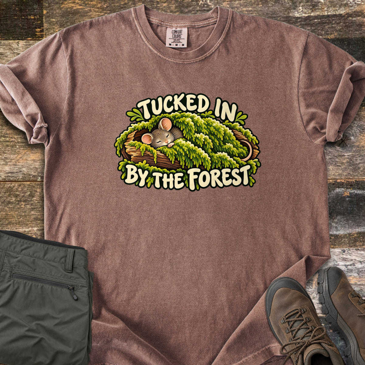 Tucked in by the Forest Comfort Colors T-shirt in Espresso, laid flat. A sleeping cartoon mouse lies under a blanket of green moss in a hollow log between the words Tucked In By The Forest. Folded olive hiking pants and brown hiking boots sit on weathered wood planks.