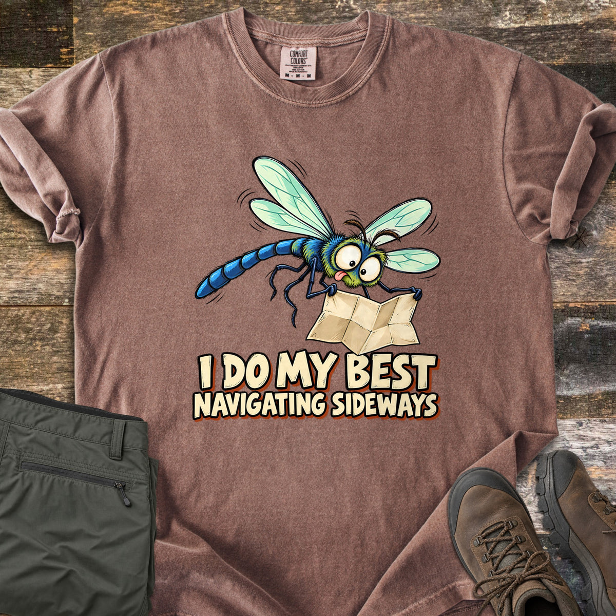 I Do My Best Navigating Sideways Comfort Colors T-shirt in Espresso, laid flat. A wide-eyed cartoon dragonfly with its tongue out holds a folded paper map above the words I Do My Best Navigating Sideways. Olive pants and brown hiking boots lie beside it on weathered wood planks.