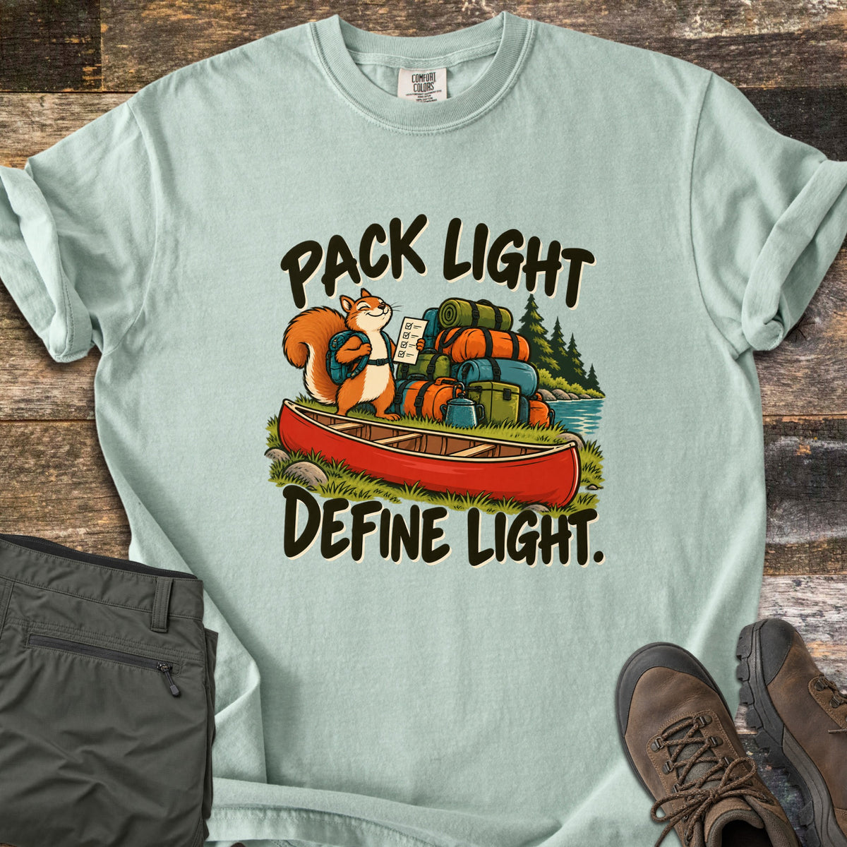Pack Light Define Light Comfort Colors T-shirt in Bay, laid flat. A smiling cartoon squirrel with a backpack holds a checklist beside a red canoe piled with gear bags, between the words Pack Light and Define Light. Olive pants and brown hiking boots sit on weathered wood.